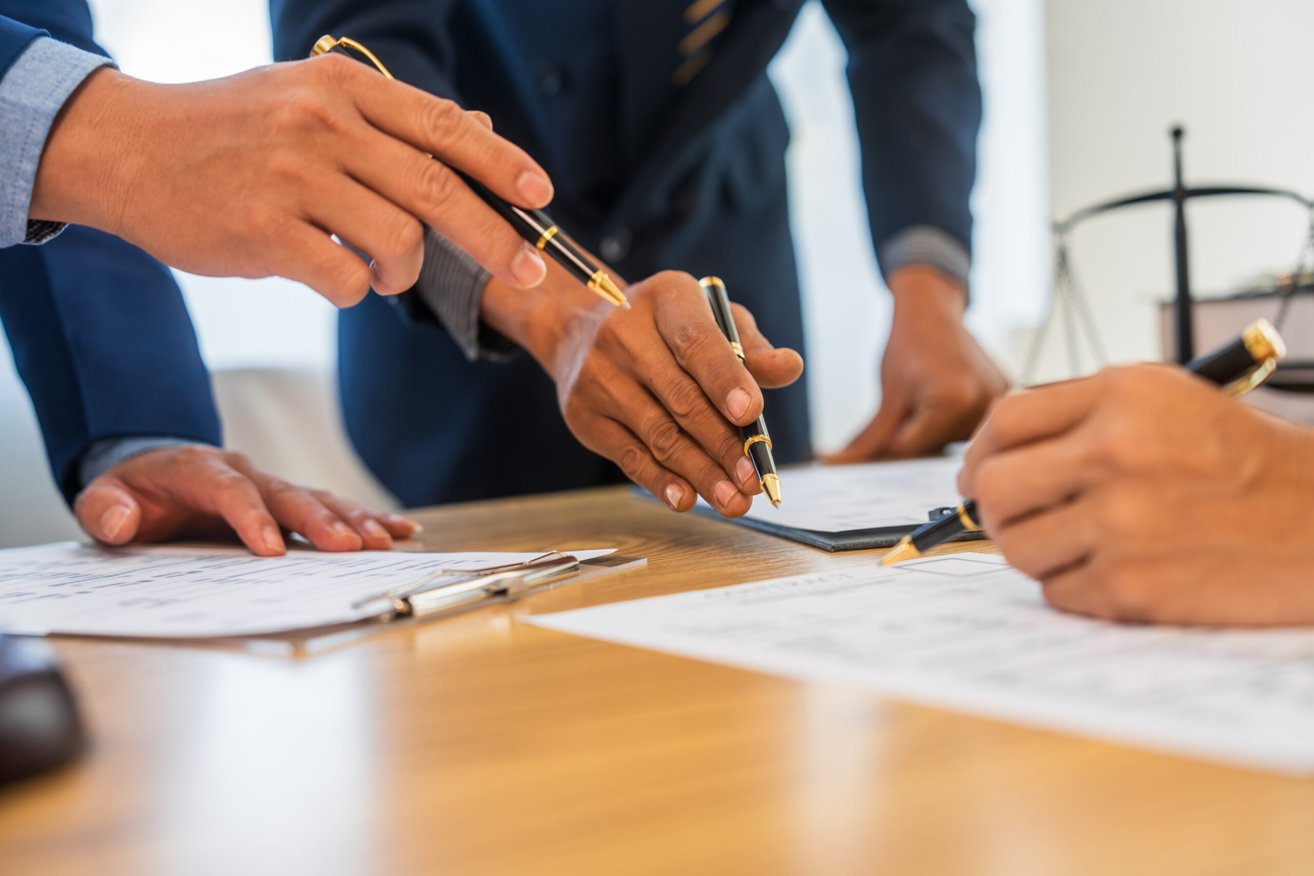 A team meeting of business people and a lawyer in formal suits is taking place at a desk, discussing a contract and various aspects of the law and litigation.